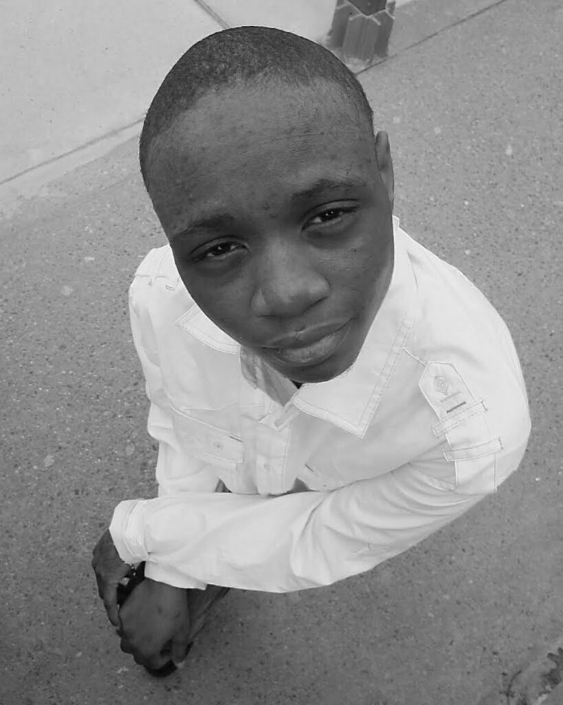 Black-and-white portrait of a masculine-presenting niiji/womyn/american indian/ indigenous individual in a structured white shirt, gazing directly into the camera with a composed and introspective expression. Shot from a high angle on a city sidewalk, their hands are clasped calmly, knees bent in a partial crouch. The image radiates grounded confidence and dignity.
