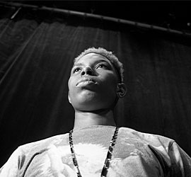 Black-and-white low-angle portrait of a masculine-presenting Black individual standing on stage, wearing a bold printed shirt and a chain necklace. Their short blond hair contrasts the dark backdrop curtain behind them. They look upward with strength and poise, symbolizing power and presence.